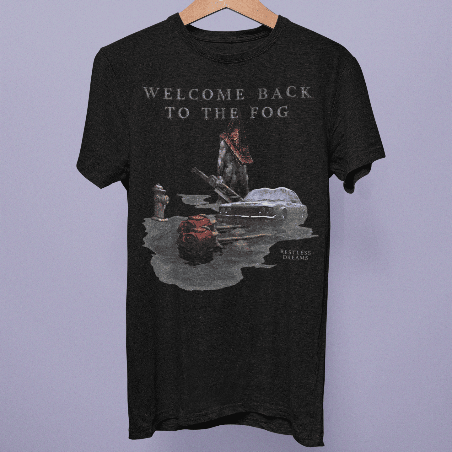 Front view of the Silent Hill Collection T-shirt on a hanger, showing “Welcome Back to the Fog” artwork with a red pyramid figure and abandoned car on black fabric.