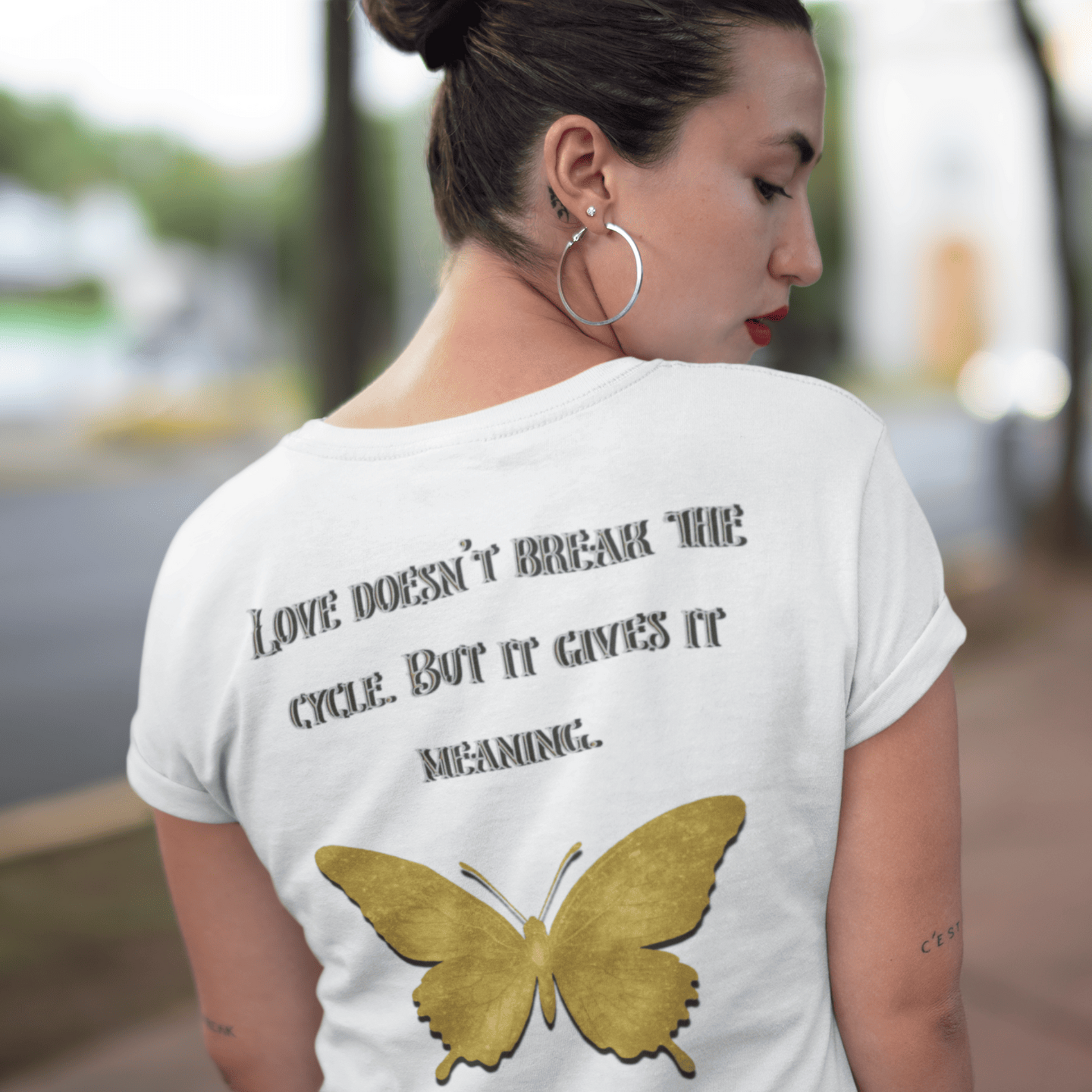 Woman wearing a white t-shirt with a gold butterfly and text on the back, standing outdoors.