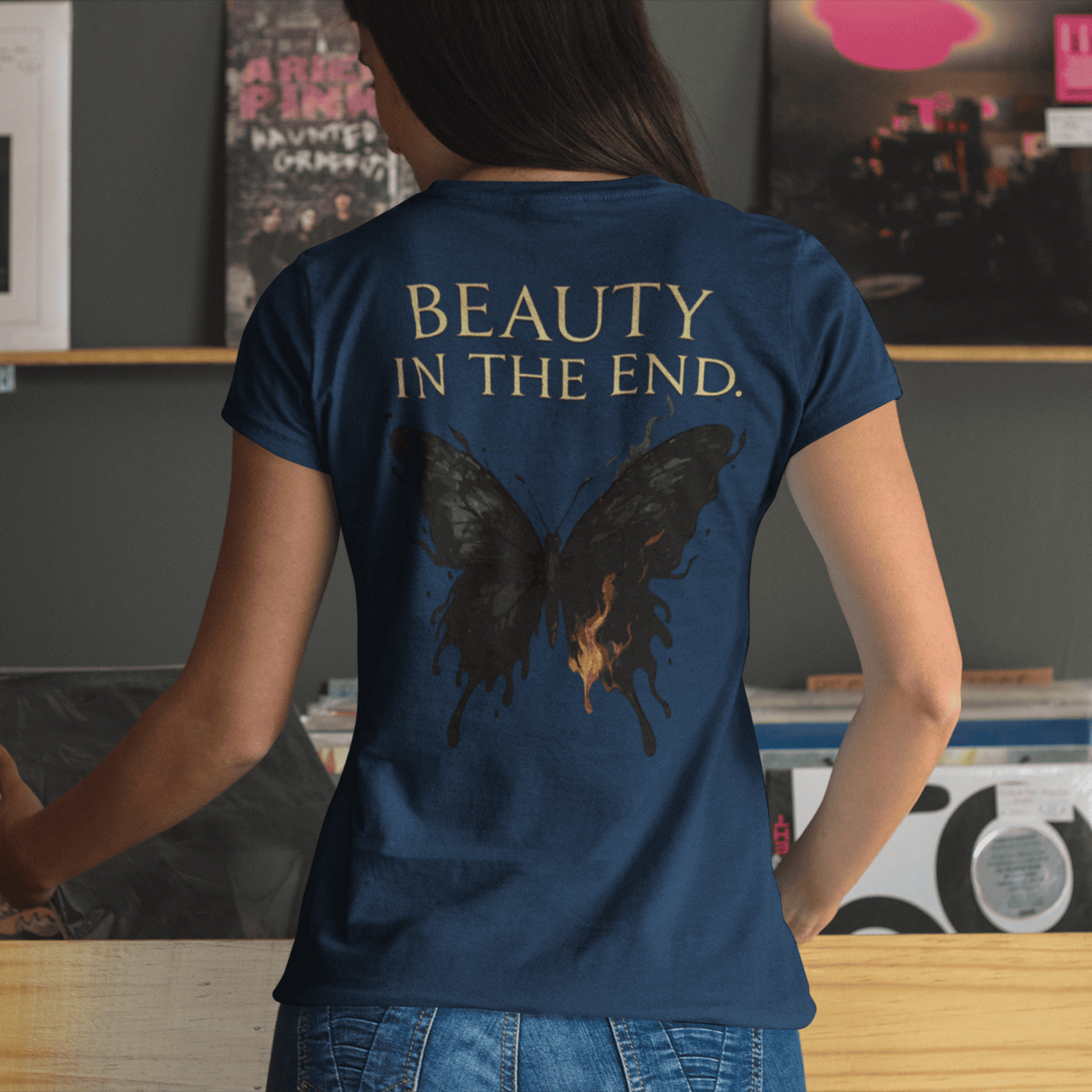 Person wearing a blue t-shirt with a butterfly design and 'BEAUTY IN THE END.' text in a room with records and photos.