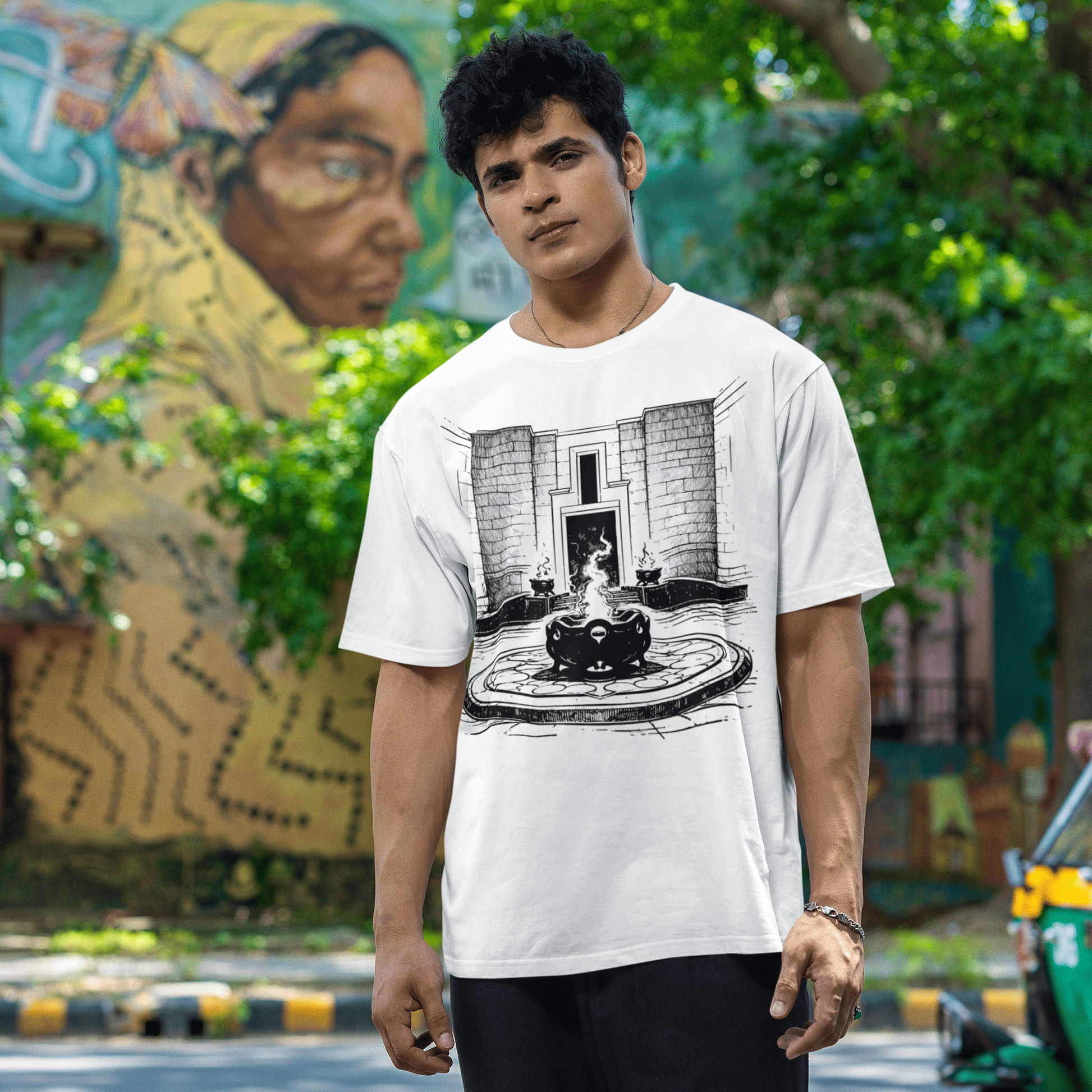 Person wearing a white t-shirt with a graphic design, standing outdoors with greenery and a mural in the background.