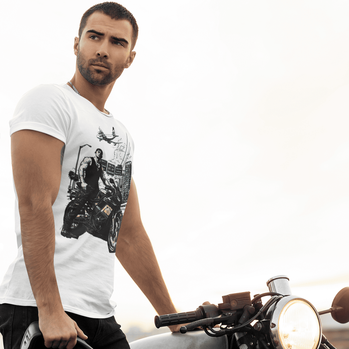 Man wearing a white t-shirt with a graphic design, standing next to a motorcycle on a beach.