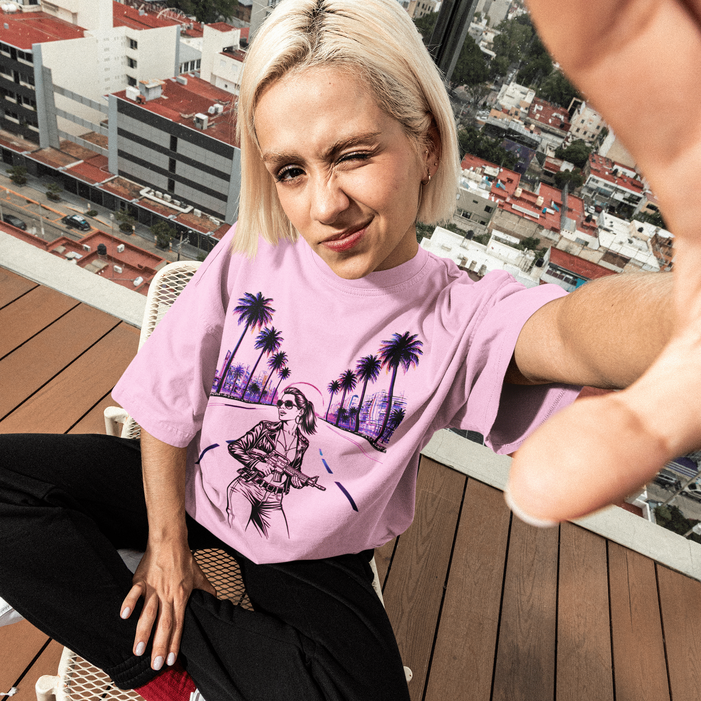 Person wearing a pink t-shirt with a graphic design, standing on a rooftop with cityscape in the background.