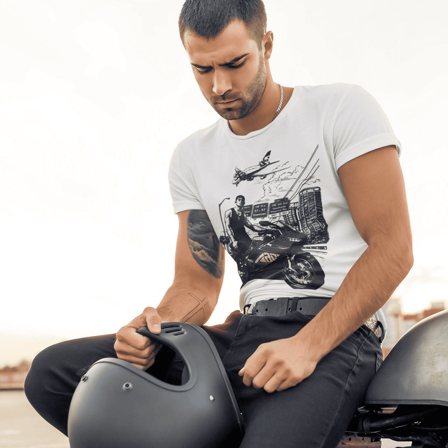 Man sitting on a motorcycle holding a helmet, wearing a white t-shirt with a graphic design.