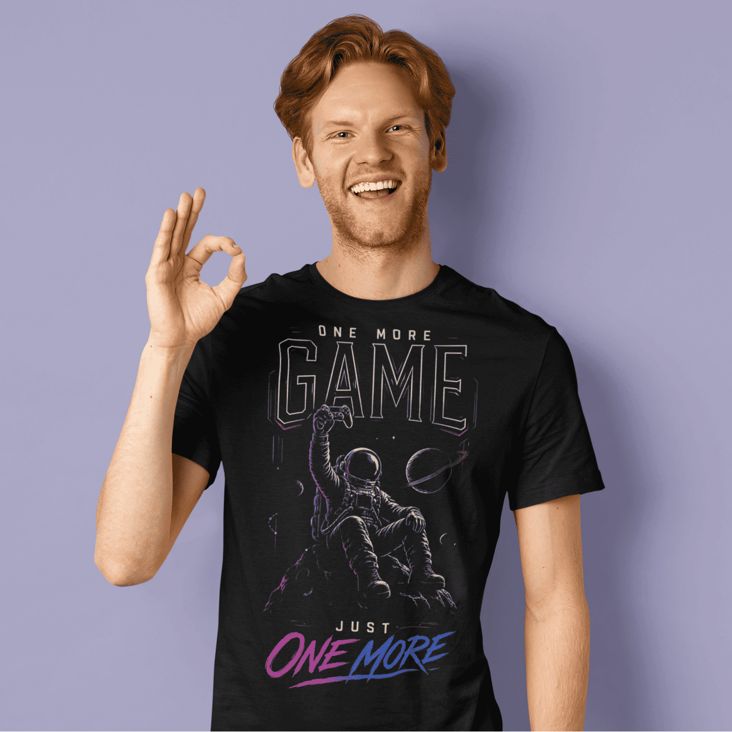 Smiling male gamer making an OK gesture in One More Game T-shirt with dark gaming artwork on purple background.