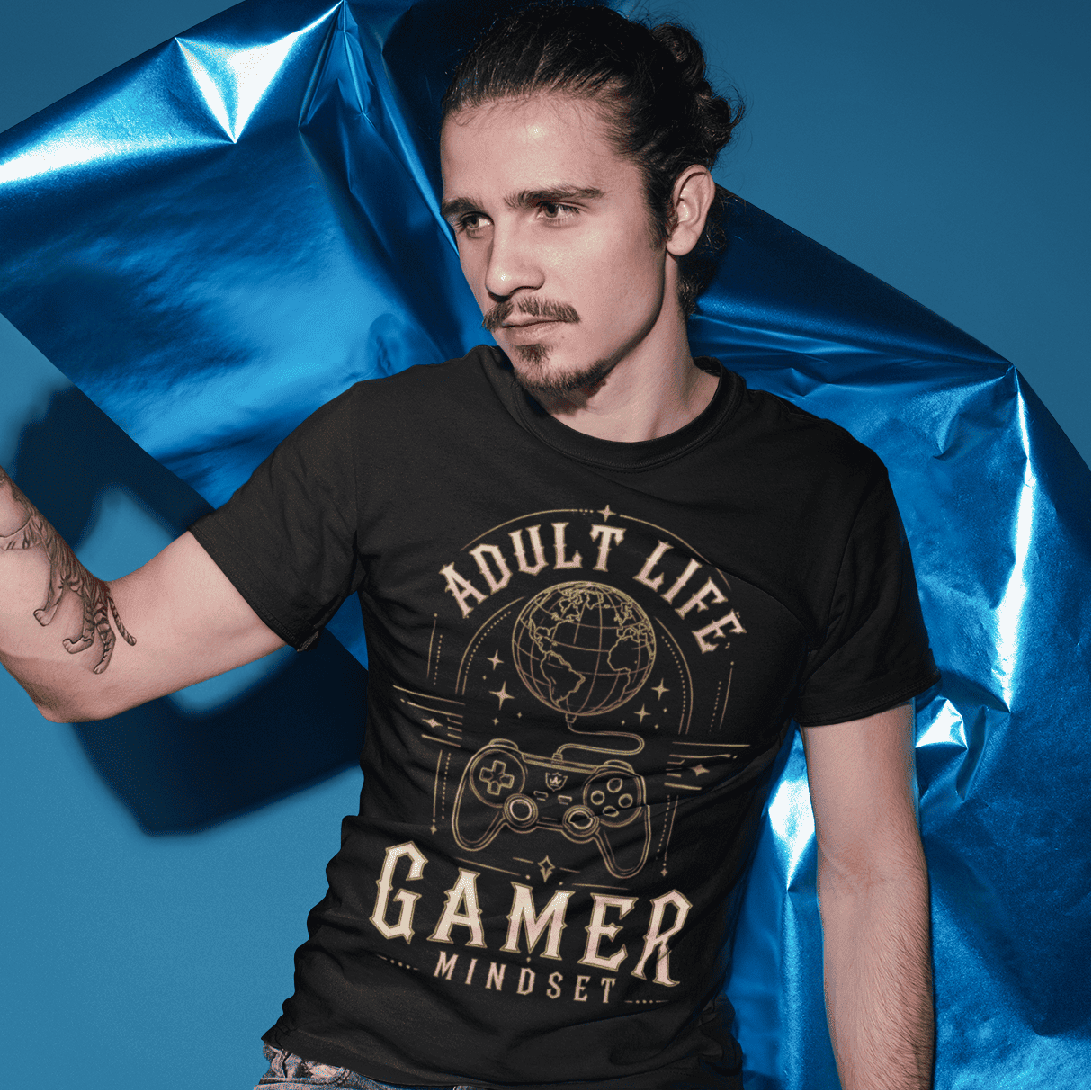 Male model wearing Adult Life Gamer Mindset T-Shirt with retro gaming graphic in a bold blue studio scene.