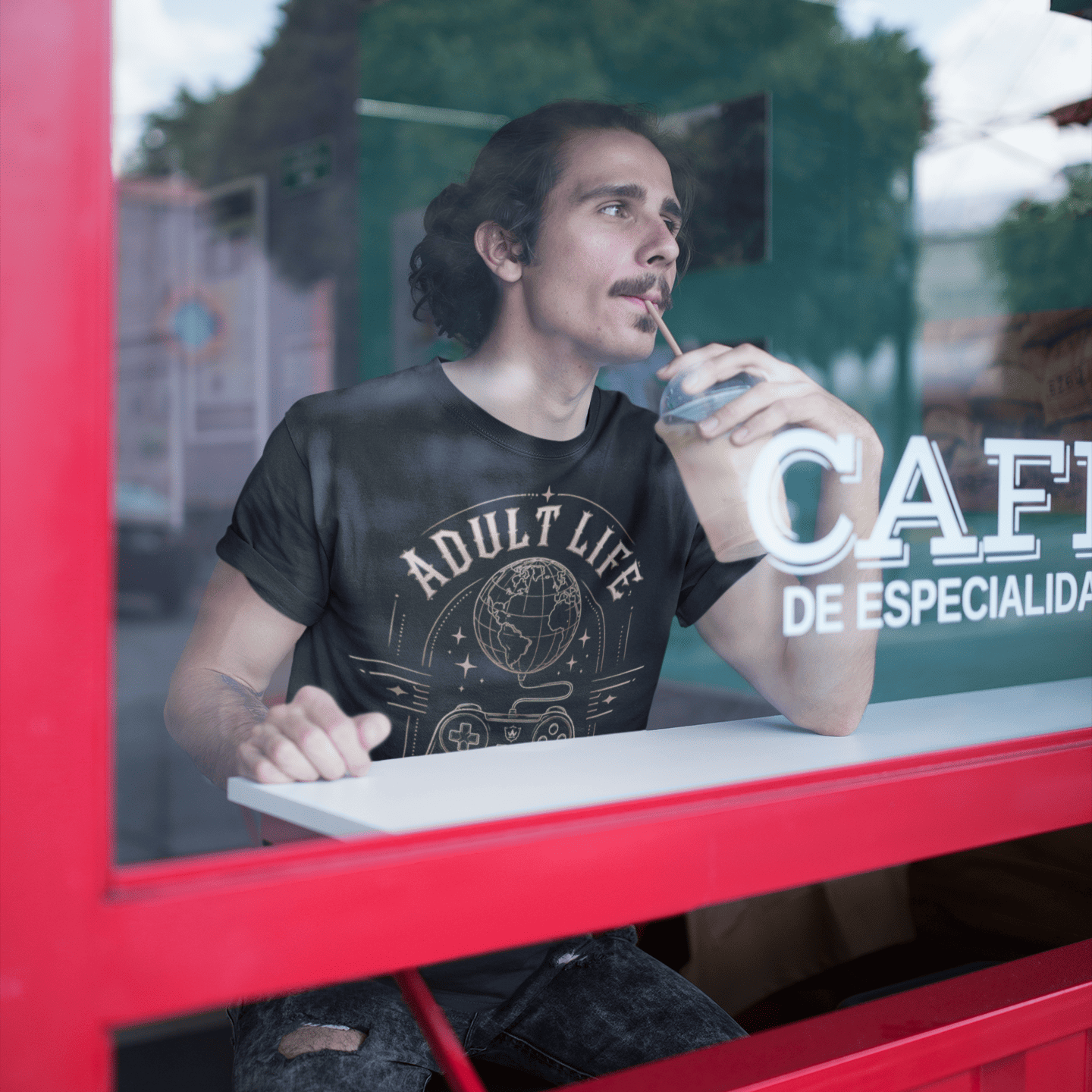 Male gamer sitting by a café window in Adult Life Gamer Mindset T-Shirt with everyday gamer mode design.